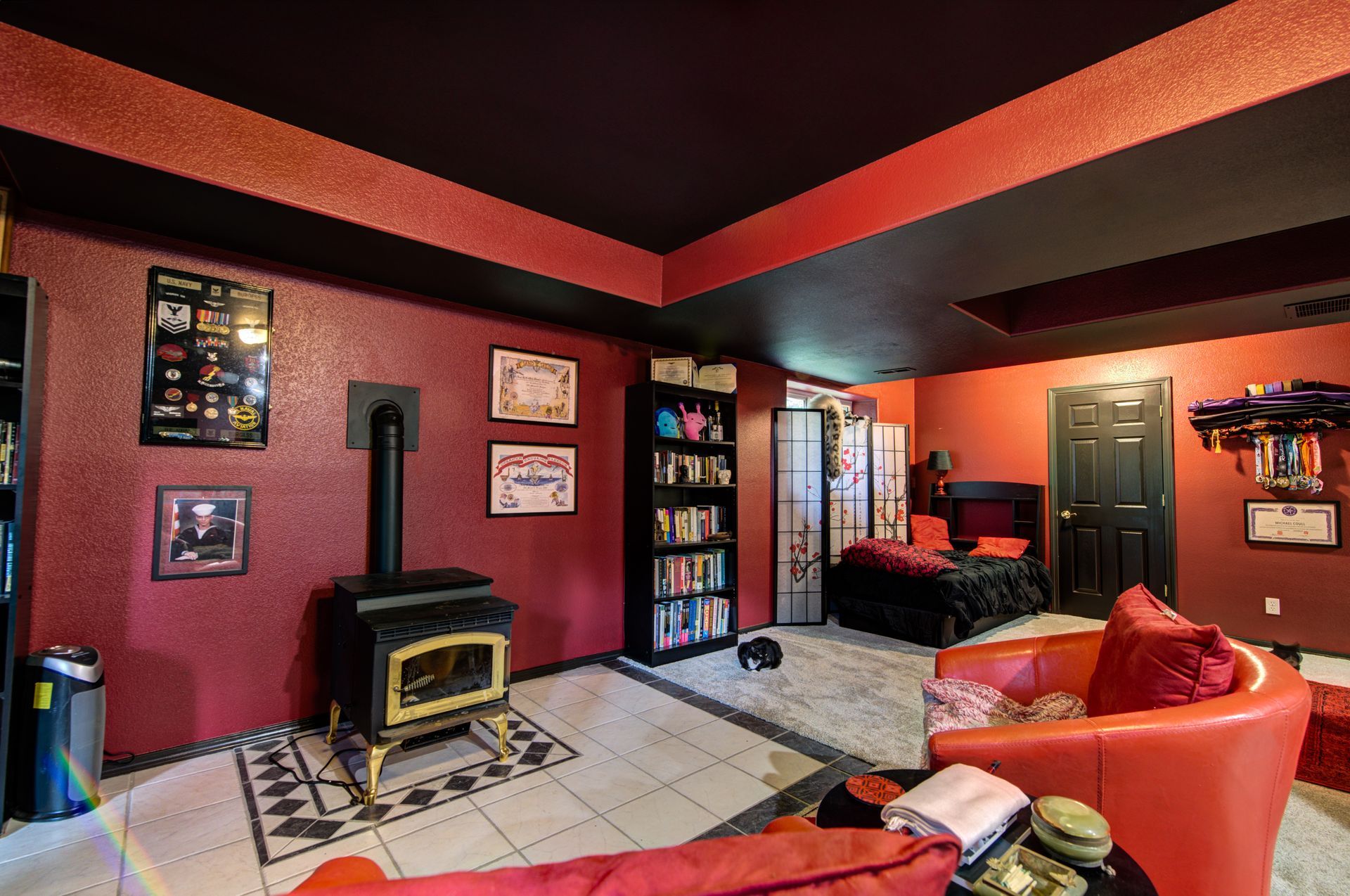 A living room with red walls , a red couch , a chair , a fireplace and a bed.