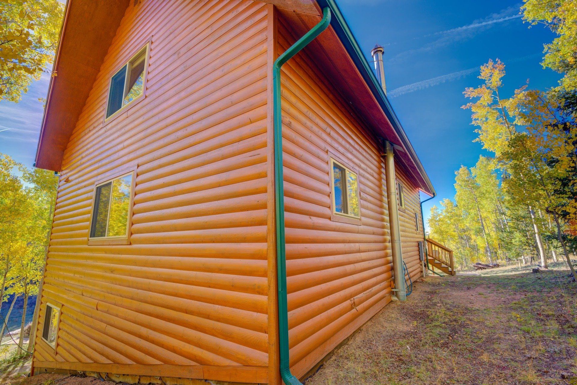 The side of a log cabin with a green gutter and trees in the background.