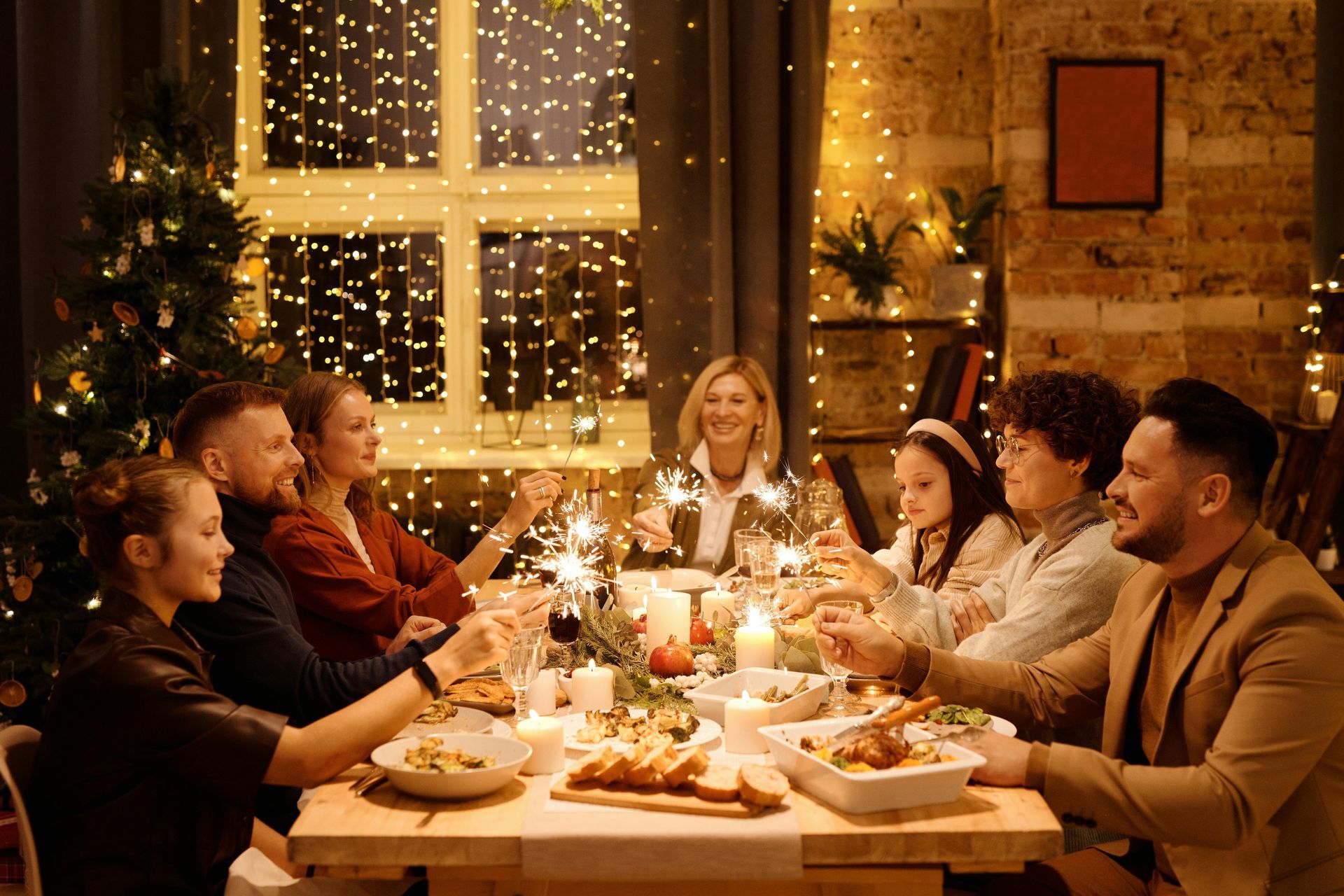 A large family is sitting at a table eating christmas dinner and holding sparklers.