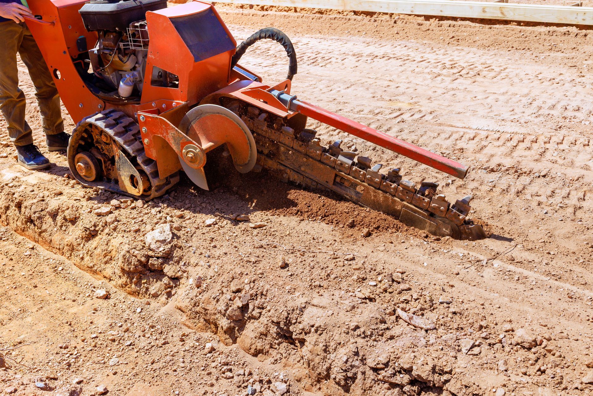 A red trencher digs into the ground of a construction site, under the sun.