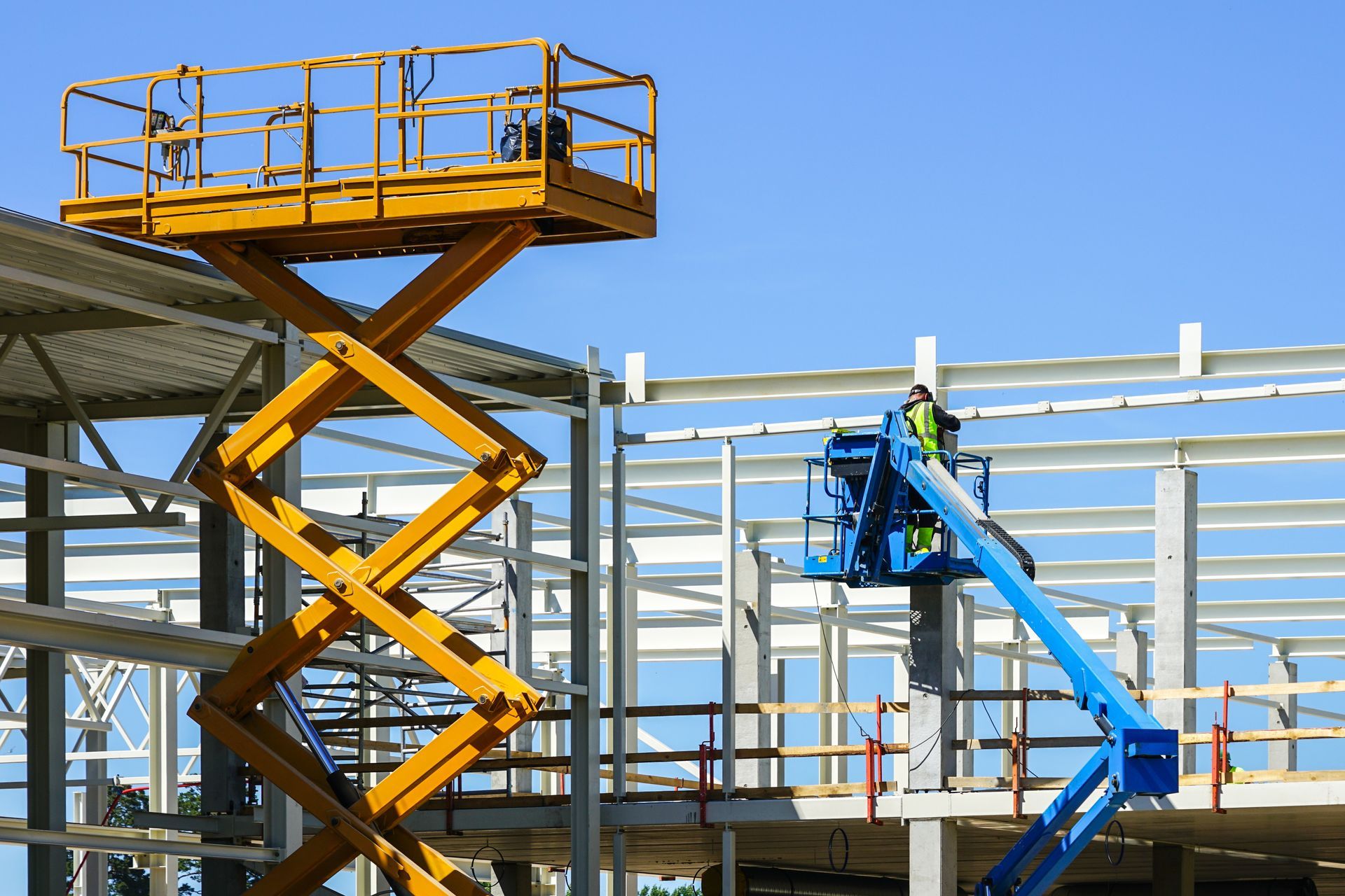 Construction site with a yellow scissor lift and a blue boom lift under a clear sky.