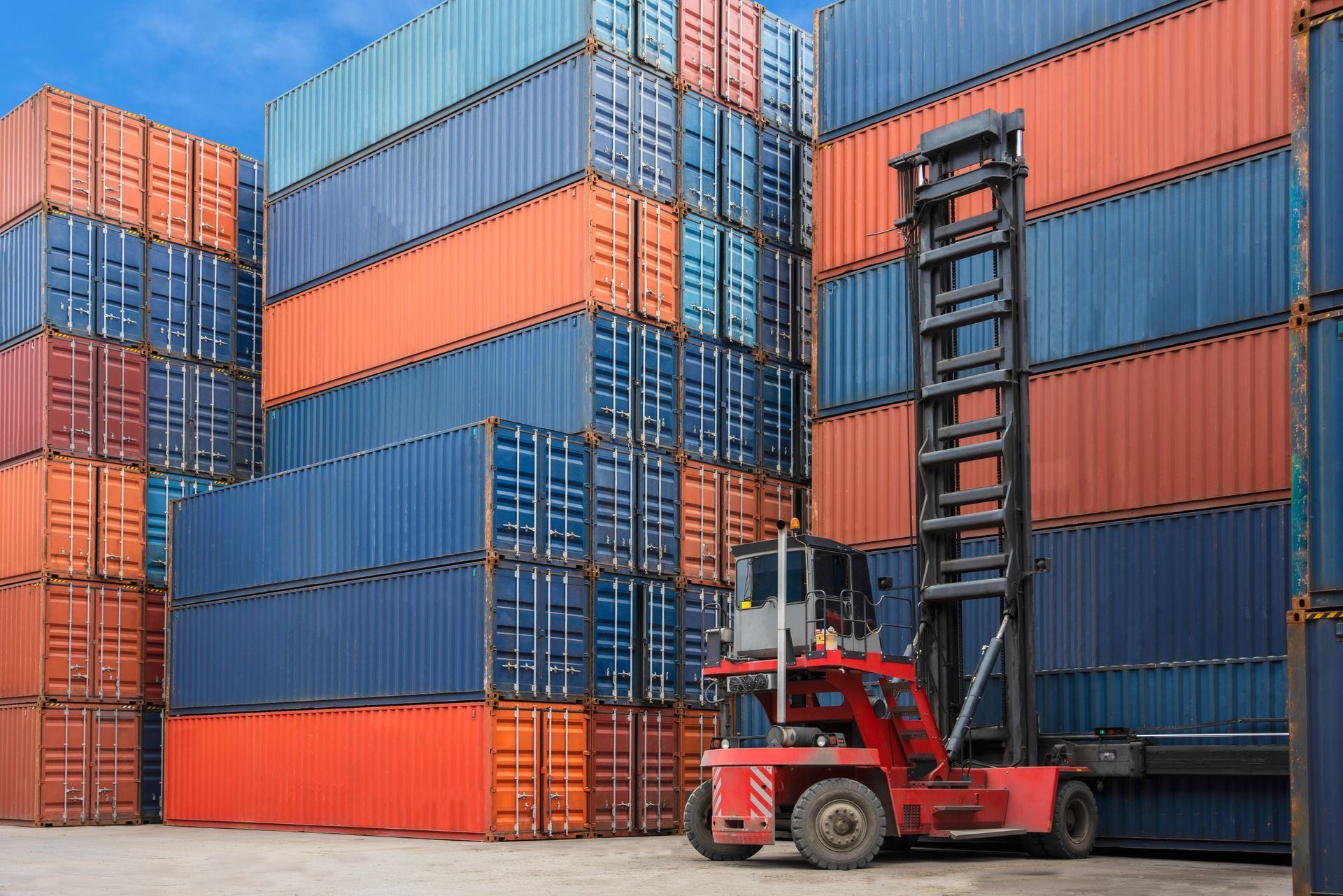 Red forklift lifts a shipping container among stacks of blue and orange containers at a port.