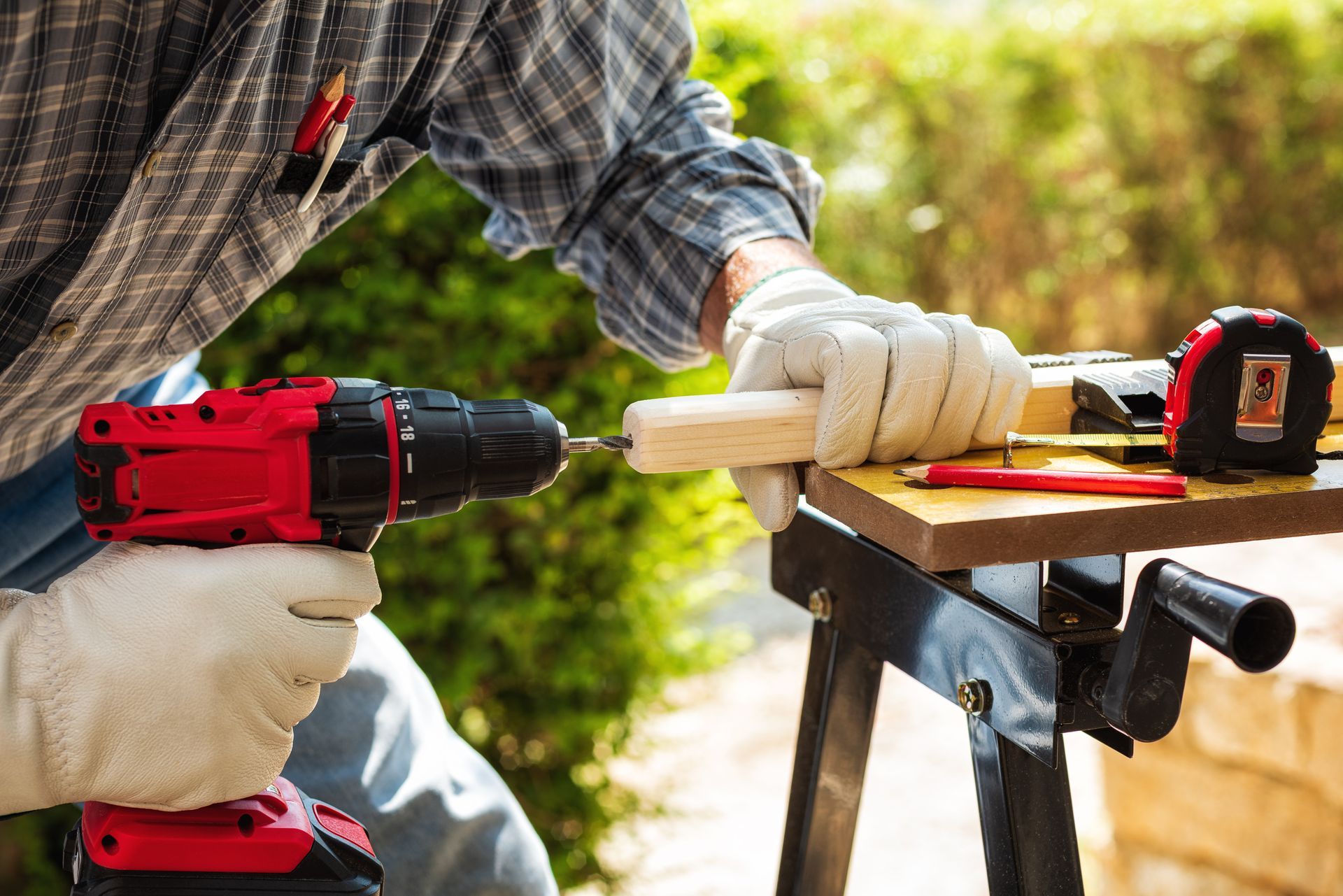 Person in gloves drilling wood with a red cordless drill on a workbench outdoors.