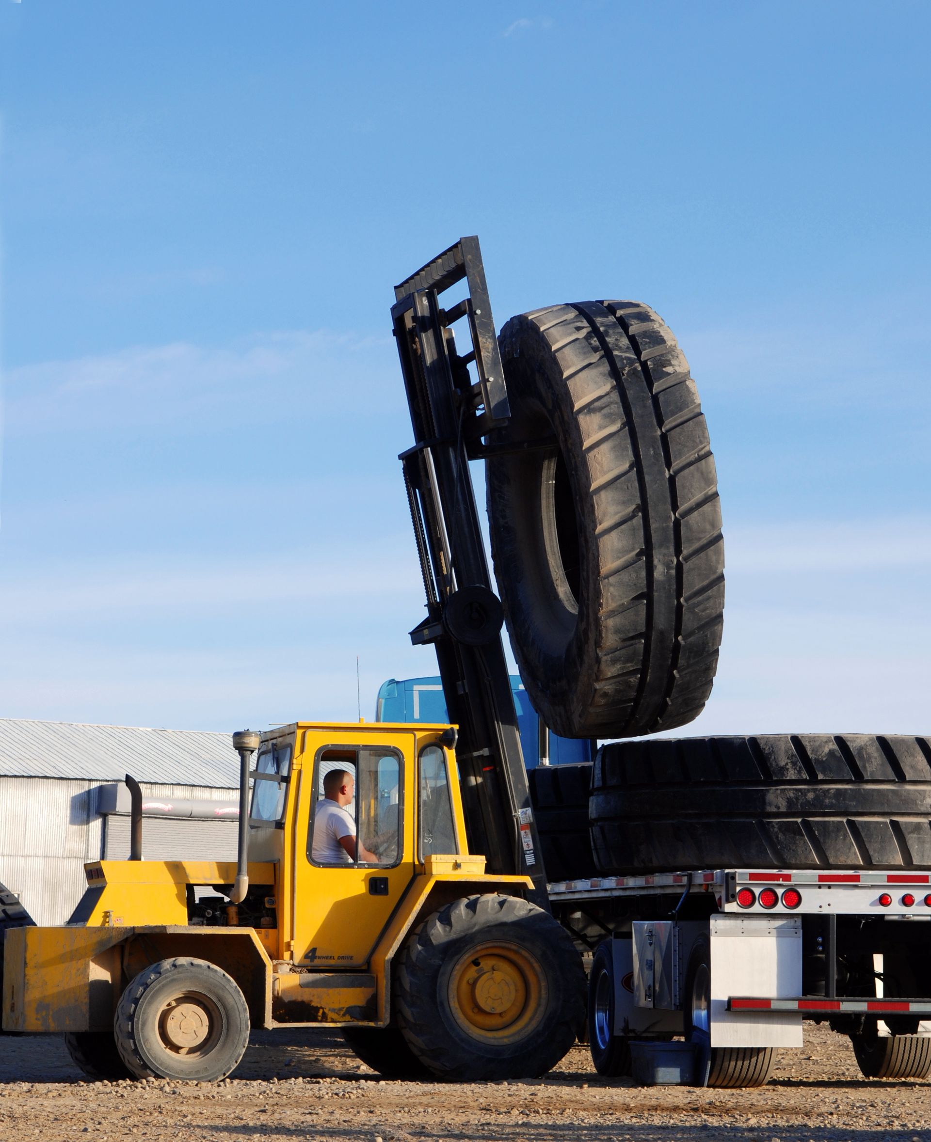 Yellow forklift lifting a large tire onto a trailer with other tires, under a blue sky.