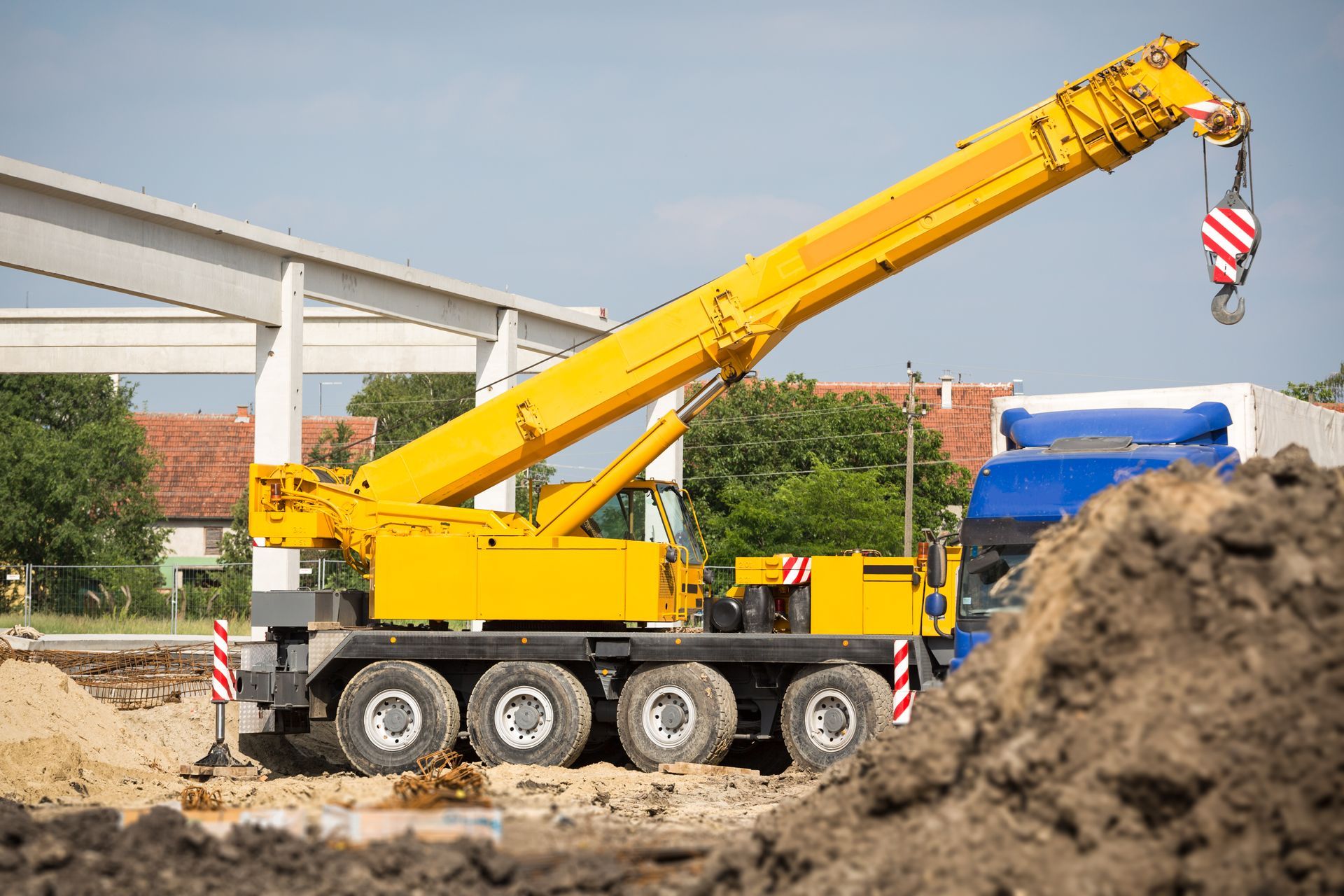 Yellow mobile crane at a construction site, lifting a load near buildings and dirt pile.