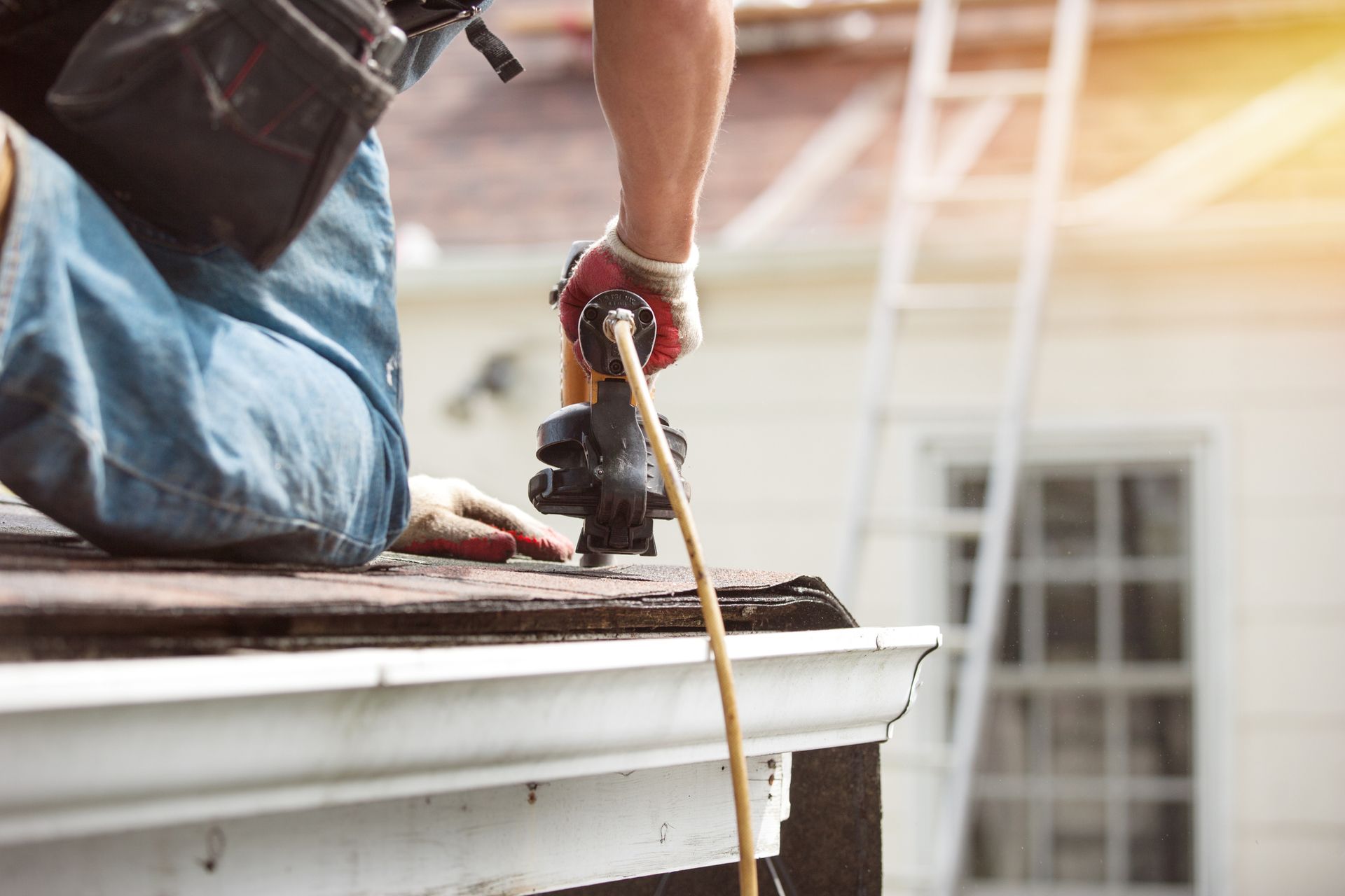 Roofer using a nail gun to install shingles on a roof near the gutter. Bright sunlight.