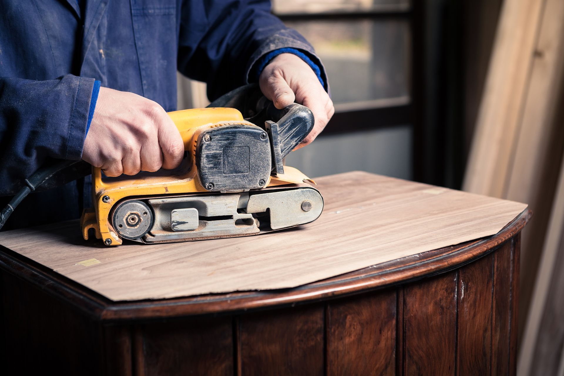 A person sanding a wooden cabinet with a yellow belt sander in a workshop.