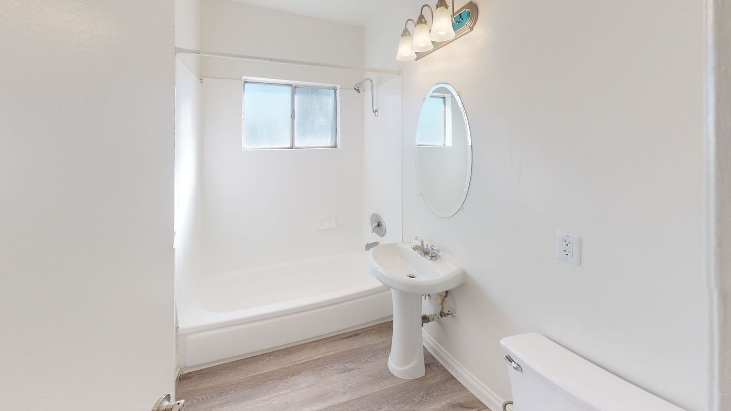White bathroom with shower/tub, pedestal sink, oval mirror, and toilet.
