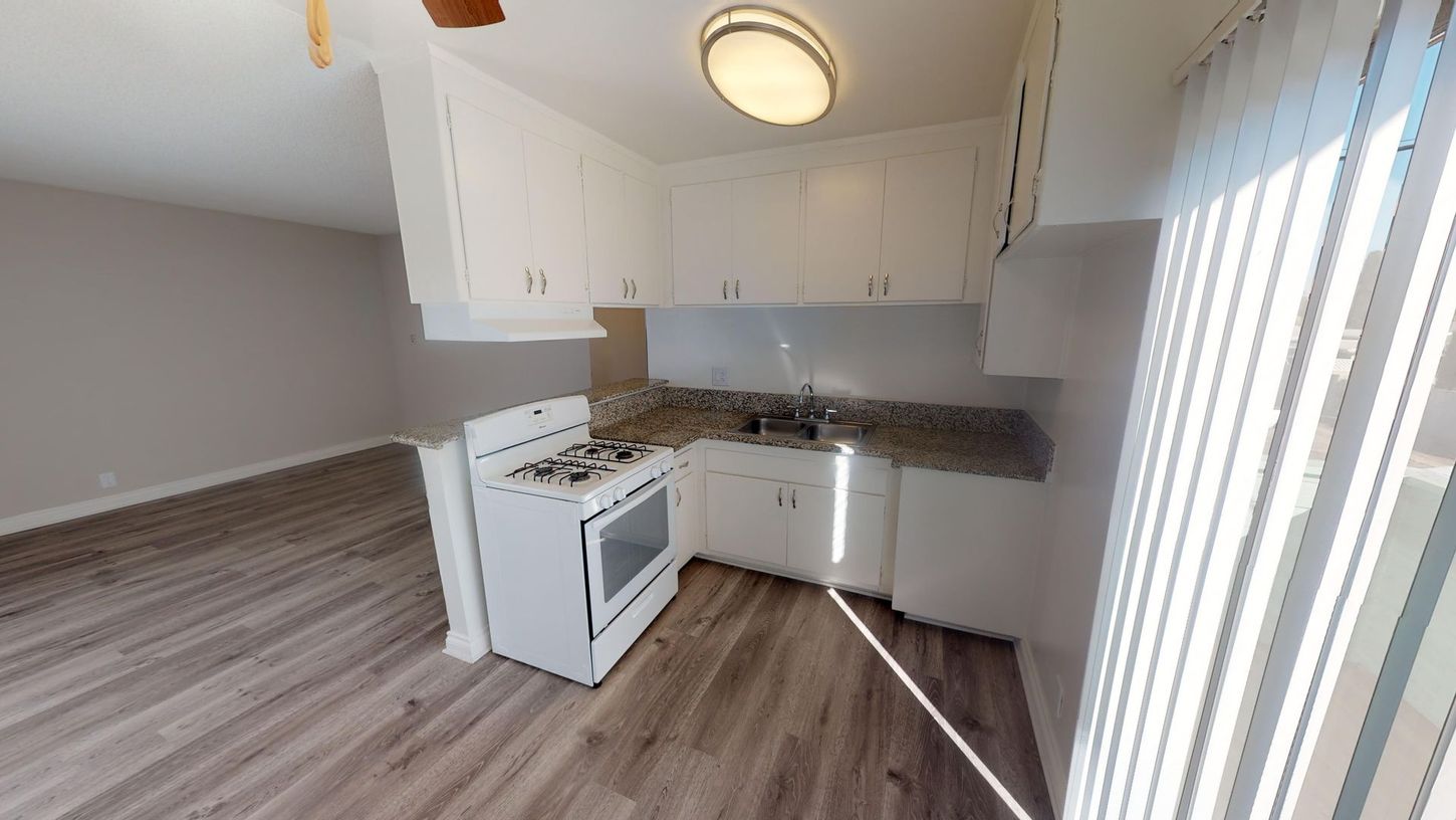Kitchen with white cabinets, stove, and light-colored flooring. Natural light streams in through vertical blinds.