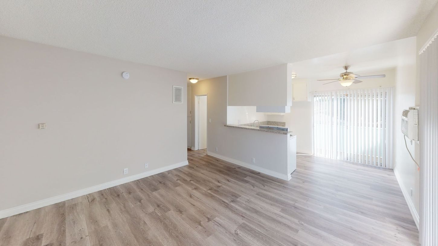 Interior view of a light-filled apartment with wood-look flooring, white walls, and a partial kitchen.