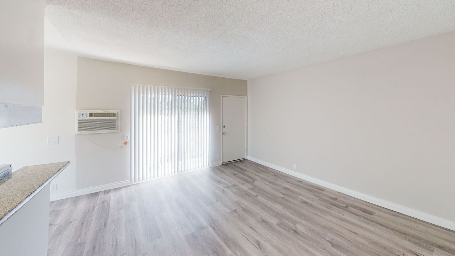 Empty living room with light grey wood-look floors, sliding glass door, white walls, and air conditioner.