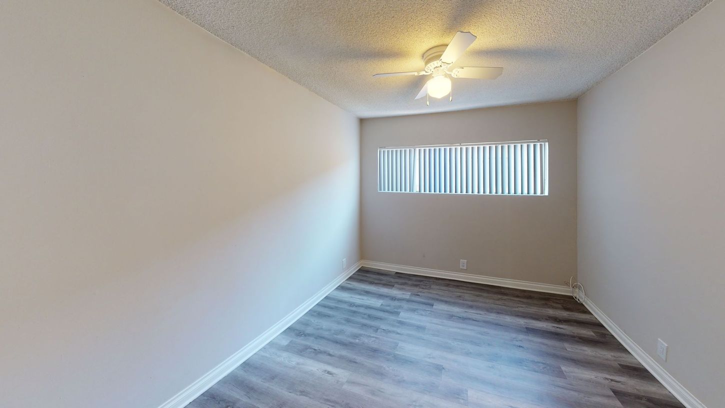 Empty room with gray laminate flooring, white walls, window with blinds, and ceiling fan.