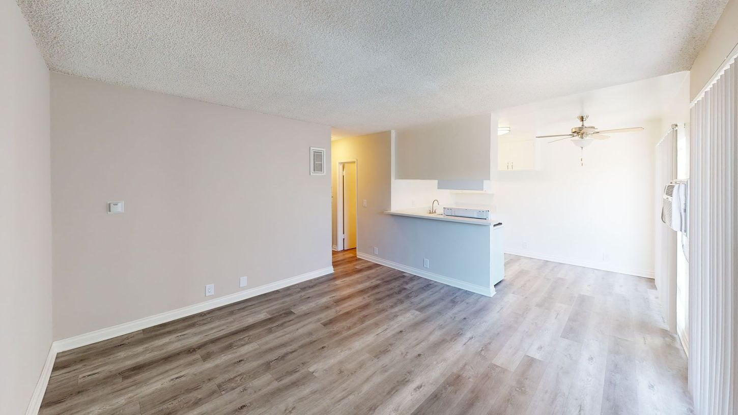 Empty apartment interior with light gray walls, wood flooring, and a small kitchen.