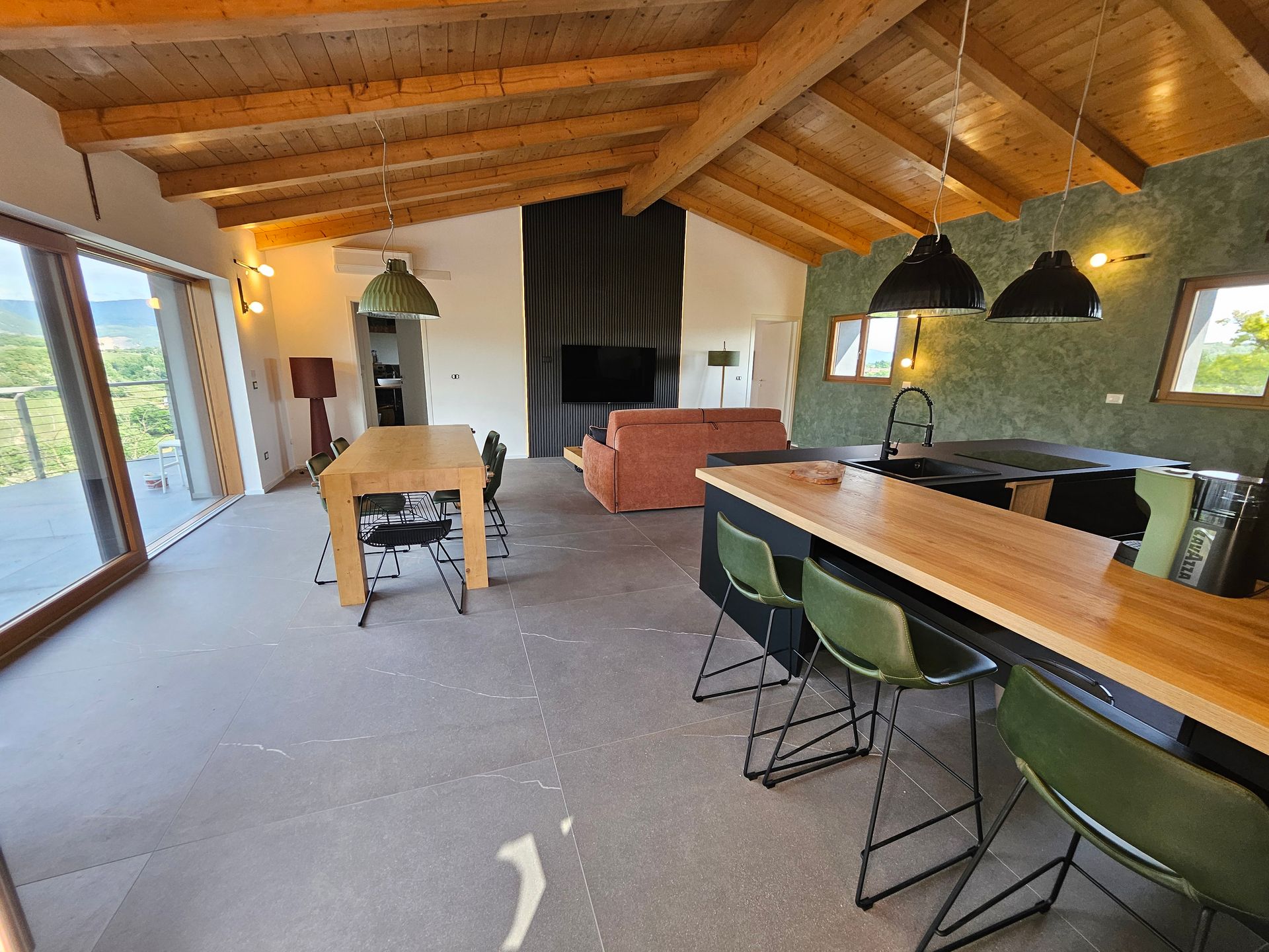 Zona pranzo in stile loft moderno con travi in legno a vista, lungo tavolo, sgabelli da bar verdi, camino e ampie finestre.