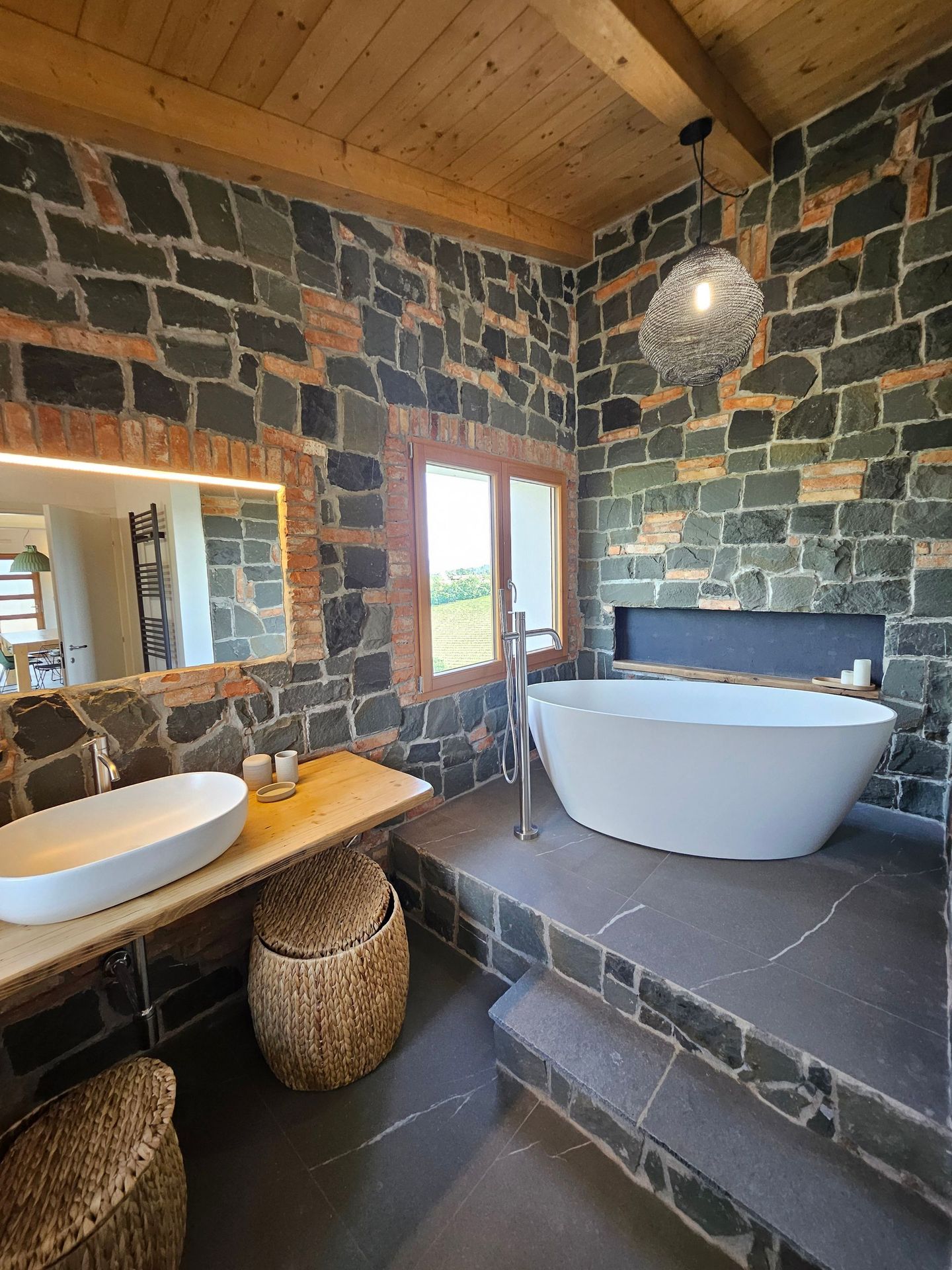 Bagno rustico con pareti in pietra, soffitto in legno, doppio lavabo e vasca da bagno freestanding su piattaforma rialzata.