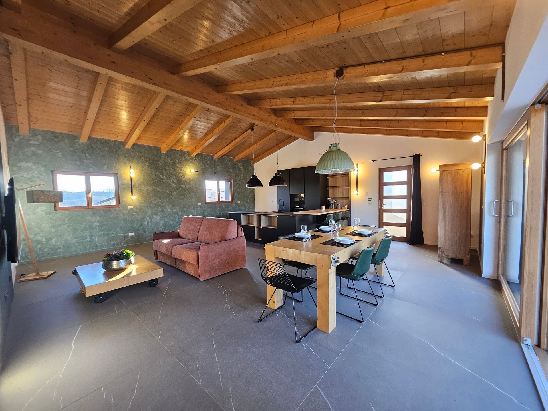 Soggiorno rustico a pianta aperta con soffitto con travi in legno, divano, tavolo da pranzo, camino e ampie finestre.