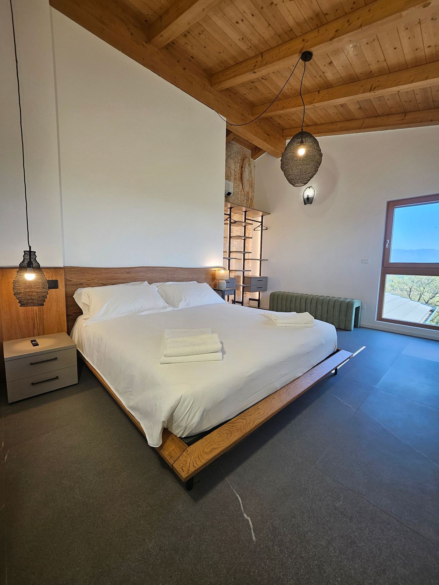 Accogliente camera da letto con letto bianco, soffitto in legno, lampada a sospensione e grande finestra con vista sull'esterno.