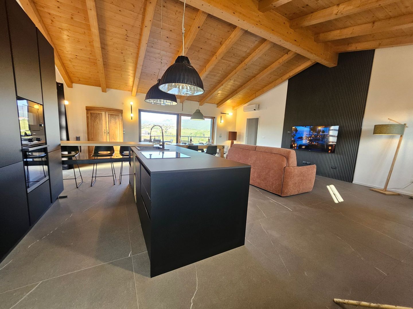 Cucina e soggiorno moderni a pianta aperta con soffitto in legno, isola nera e divano rosa vicino alle grandi finestre.