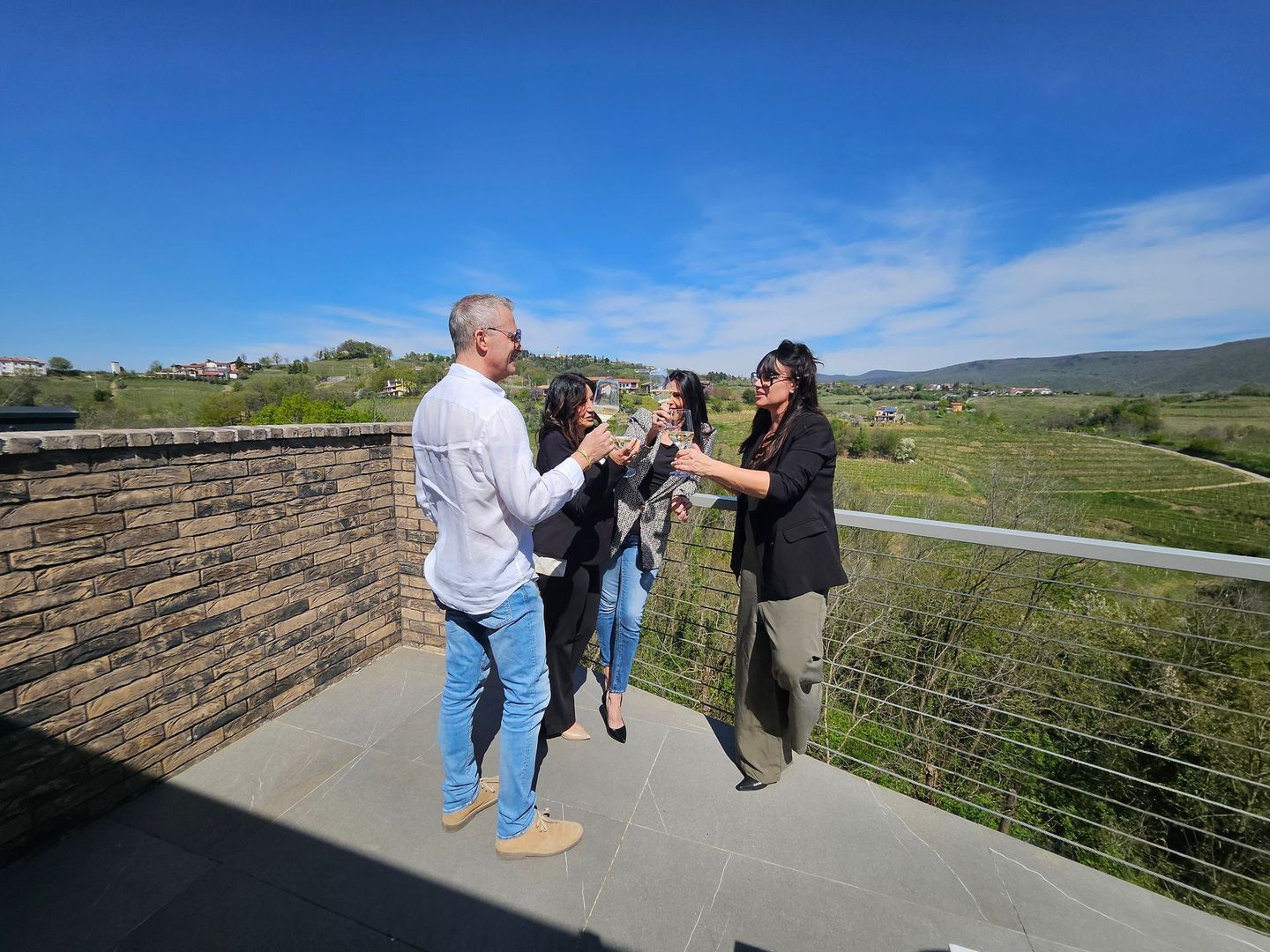 Tre persone chiacchierano su un punto panoramico vicino a un muro di pietra, con le colline e il cielo azzurro sullo sfondo.