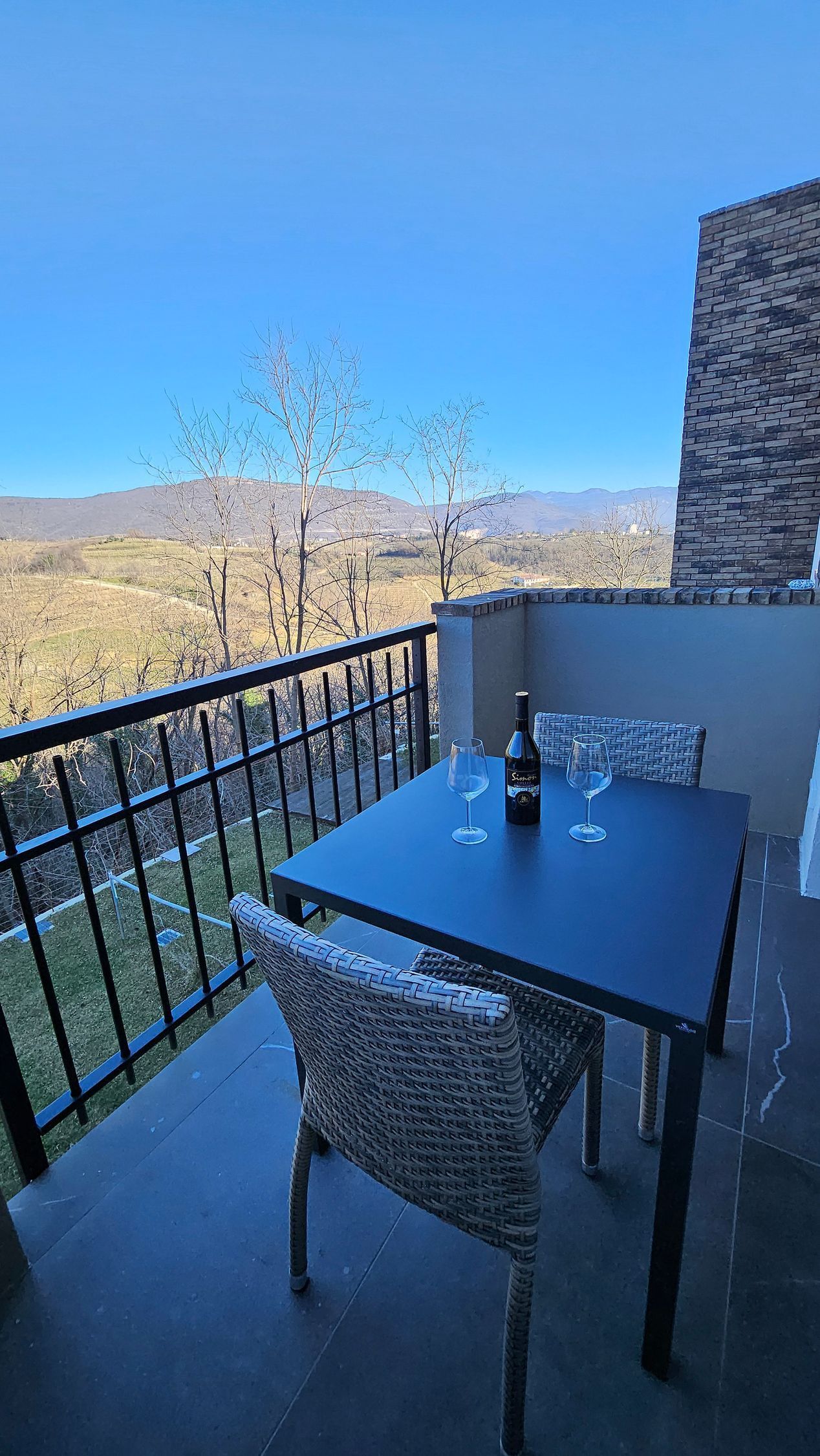 Balcone con tavolo e sedie neri, con vista su alberi e montagne sotto un cielo azzurro limpido.