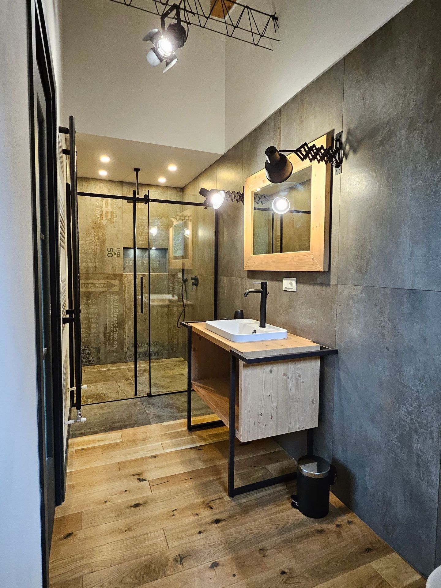 Bagno moderno con doccia in vetro, mobile lavabo in legno e specchio a parete in un interno stretto e dai toni grigi.