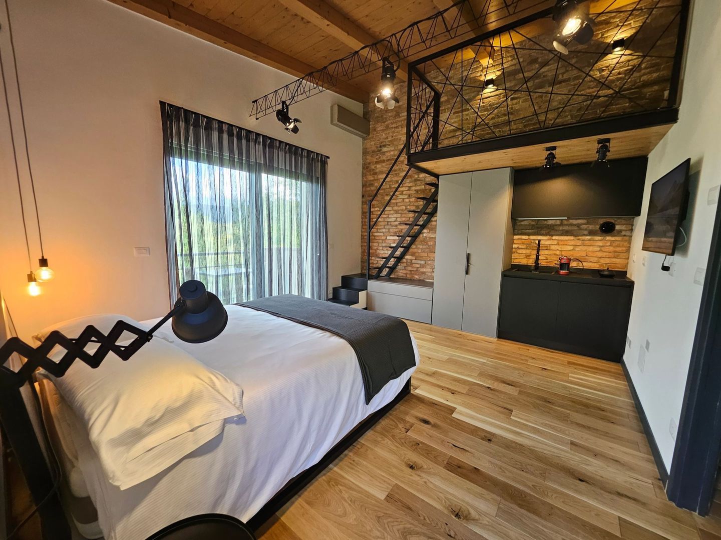 Camera da letto in stile loft moderno con soffitto in legno a vista, letto nero, ampia finestra e scala in metallo.