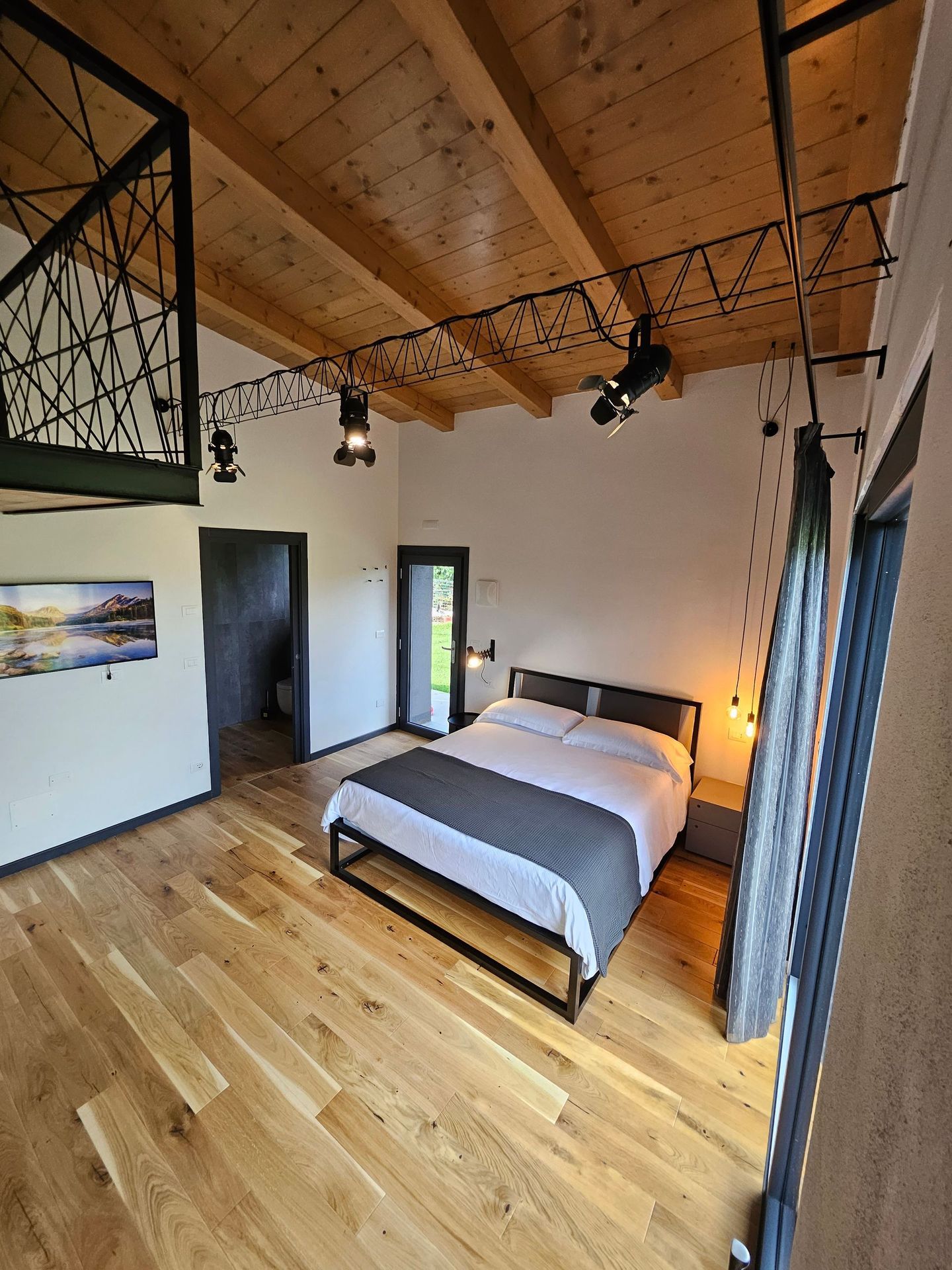 Camera da letto moderna con pavimento in legno, soffitto con travi a vista, letto, opere d'arte alle pareti e porta aperta sul balcone.