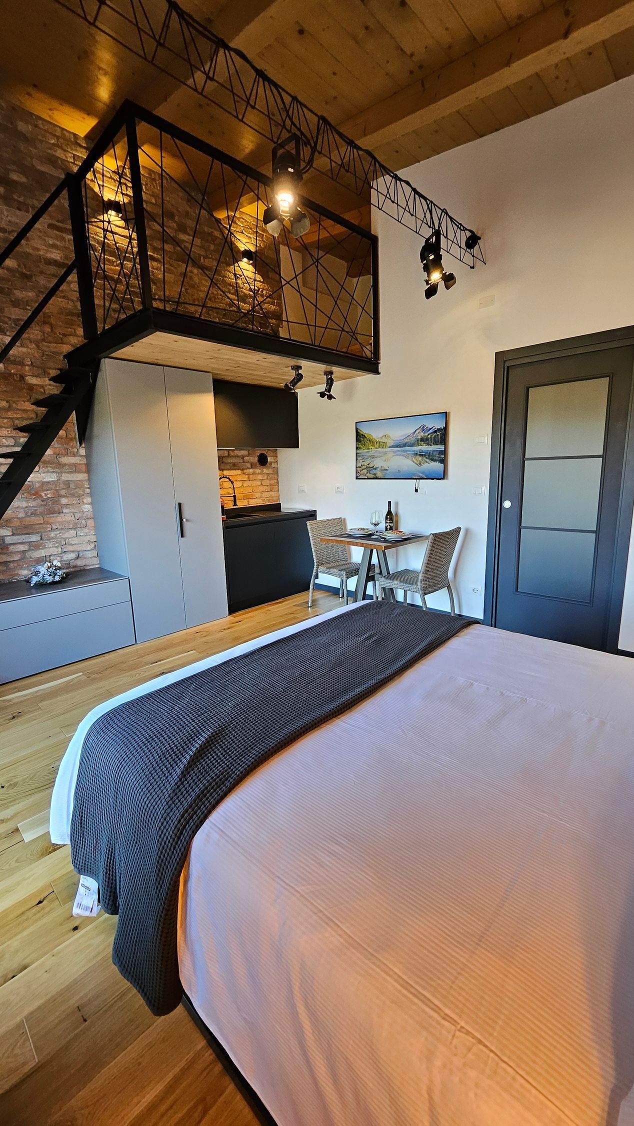 Accogliente camera da letto in stile loft con letto, parete in pietra, ringhiera in metallo nero e una piccola scrivania vicino a una finestra.