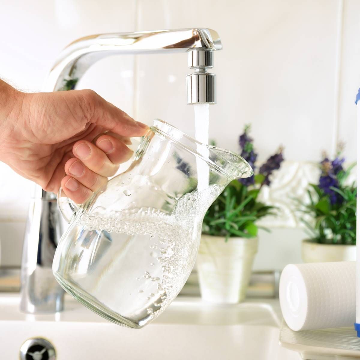 Hand filling a clear glass pitcher with water from a chrome faucet in a kitchen setting.