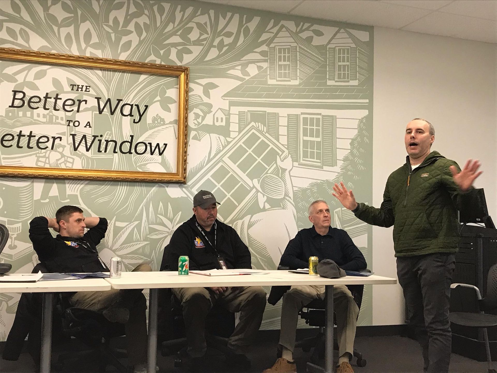 A man gestures while speaking at a table with three seated men in front of a window-themed mural.