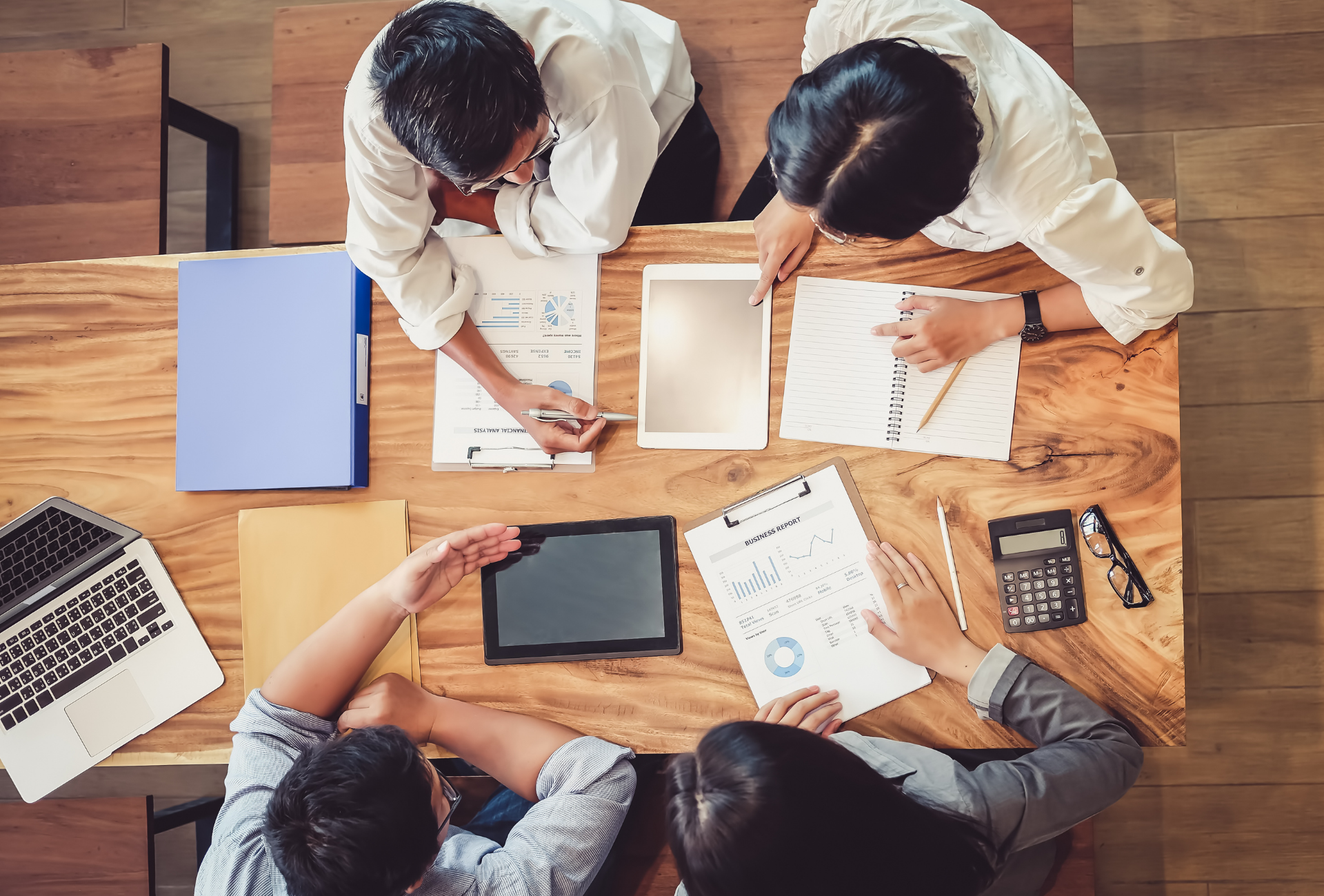 Four people at a wooden table reviewing documents, using a laptop, tablet, and calculator.