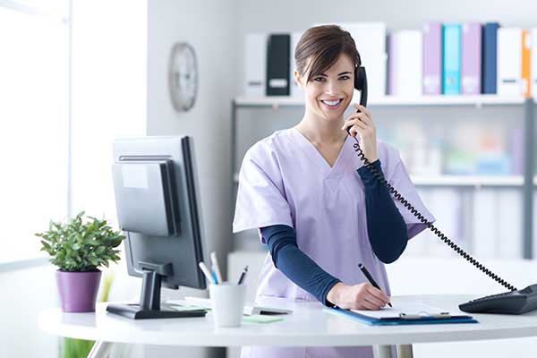 Medical office worker smiling while on the phone, writing notes at a desk.