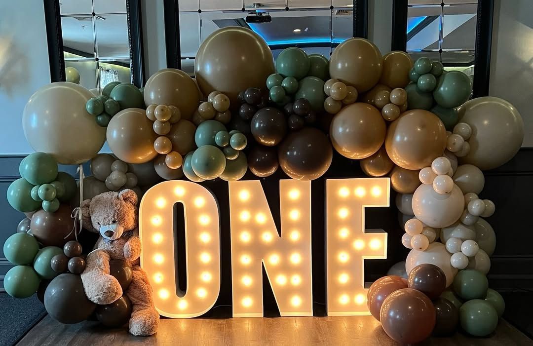 A sign that says one is surrounded by balloons and a teddy bear.