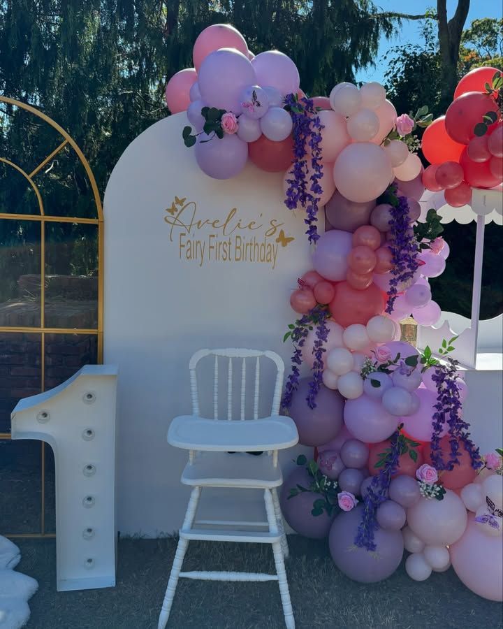A white high chair is sitting in front of a wall decorated with pink and purple balloons