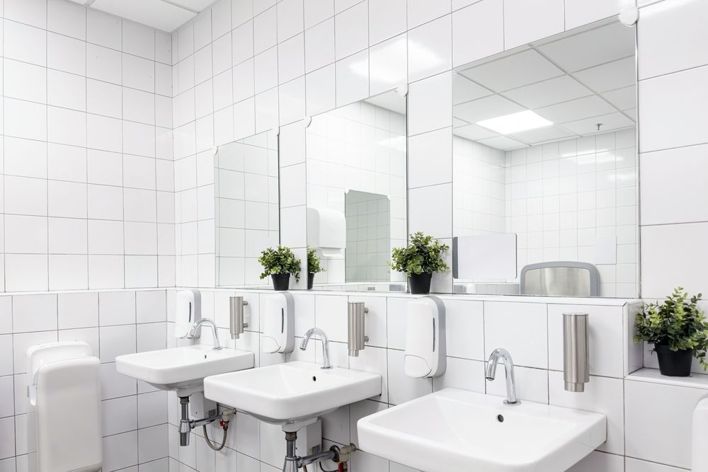 A bathroom with white tiles , sinks , mirrors and plants.