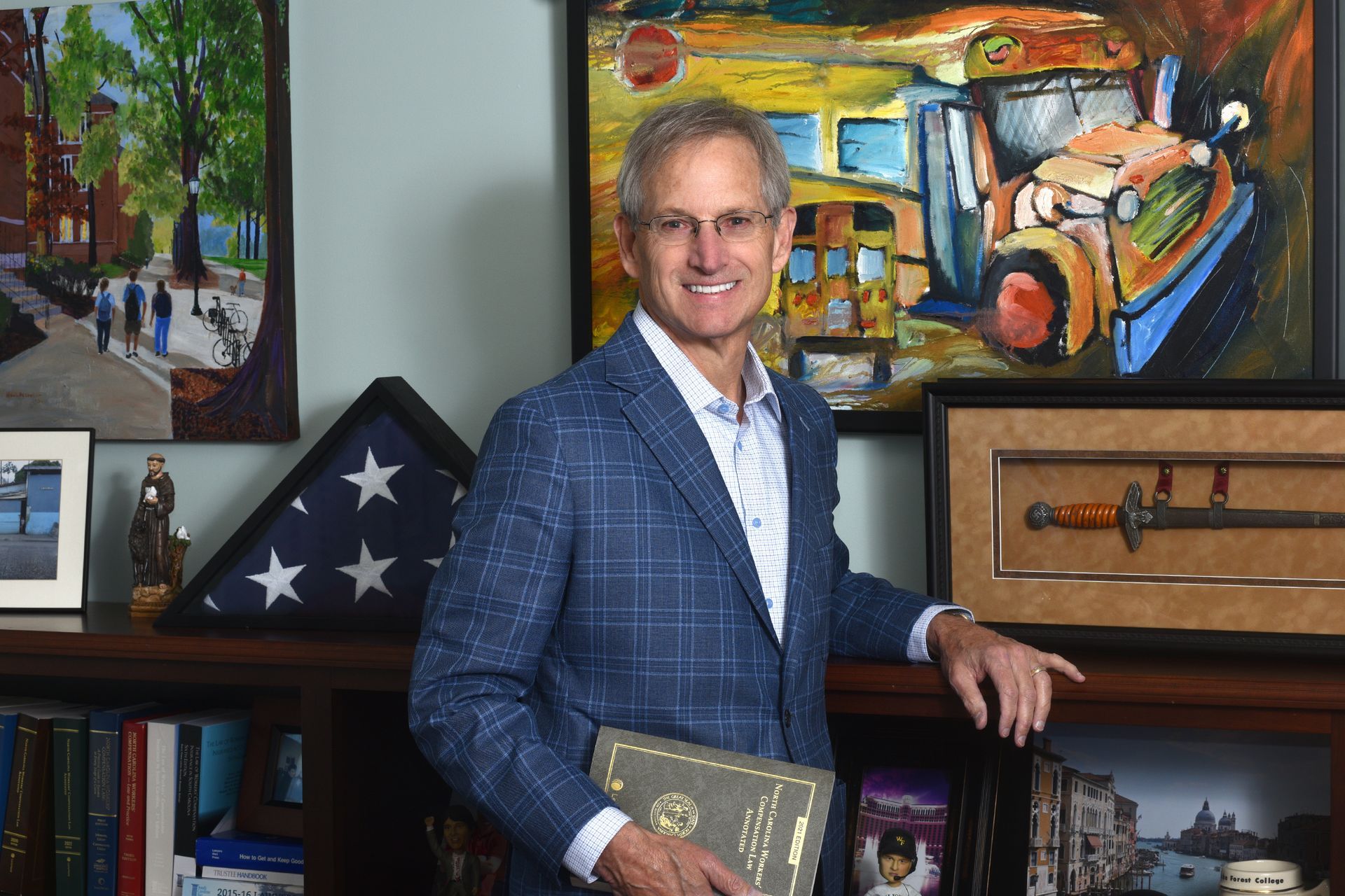 A smiling person in a blue plaid blazer poses in an office, holding a book, with paintings and decor on the shelves.