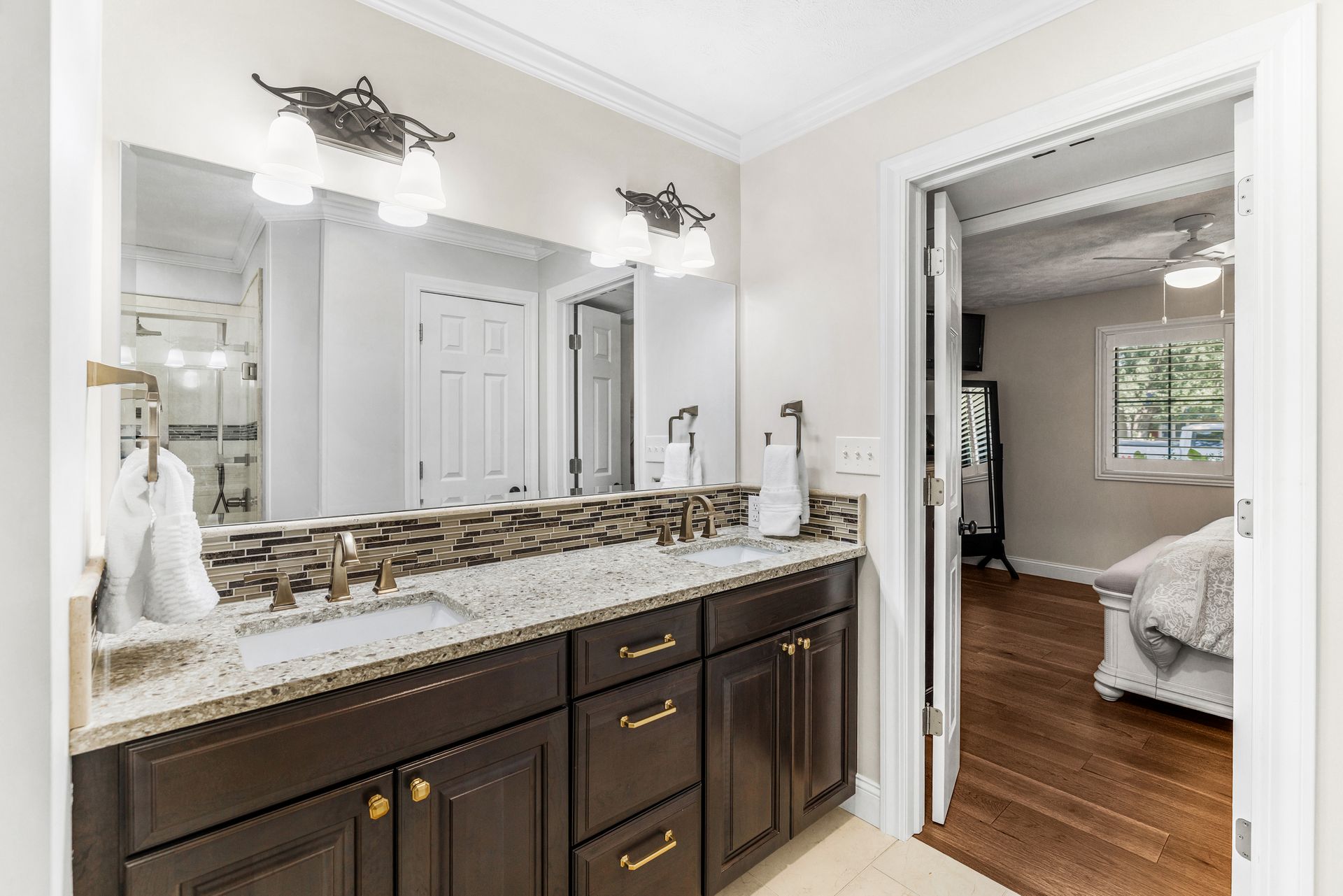 Bathroom with double sinks, brown cabinets, granite countertop, and open doorway to bedroom.