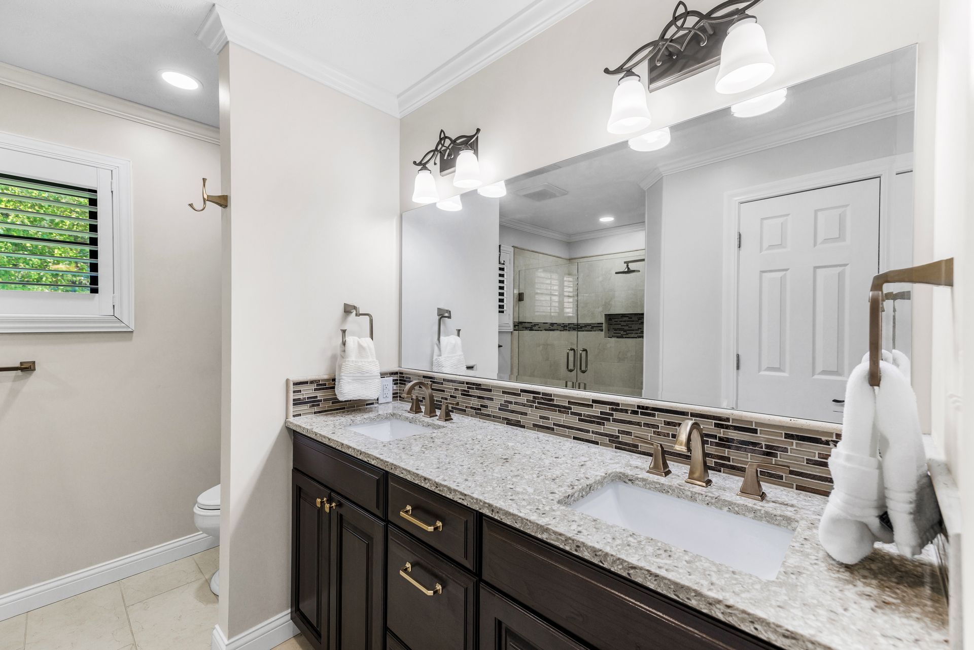 Bathroom with dual sinks, brown cabinets, granite countertop, large mirror, and decorative backsplash.