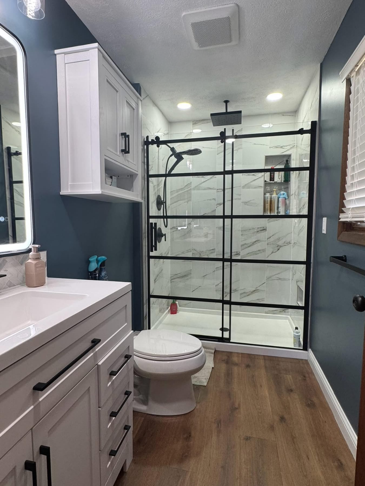 Modern bathroom with blue walls, white vanity, black-framed shower, and wood-look flooring.