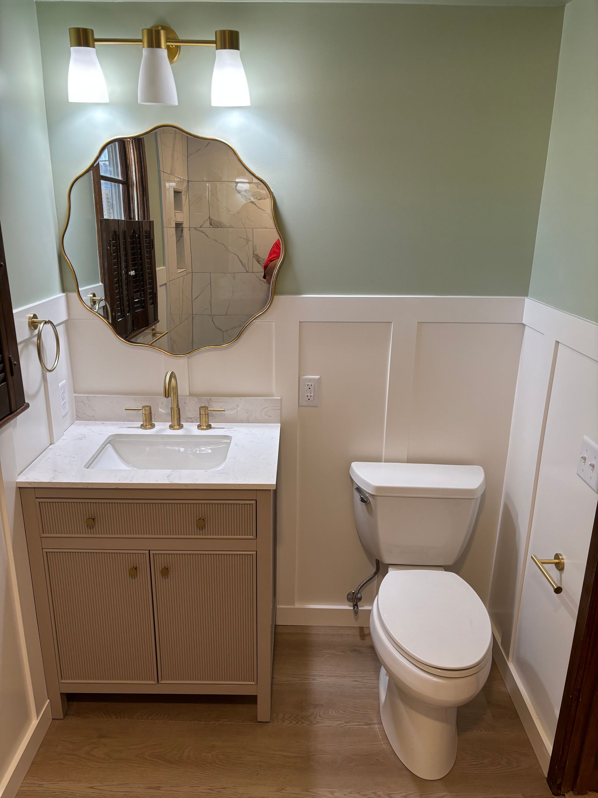 Small bathroom with vanity, toilet, and decorative mirror; sage green and white color scheme.