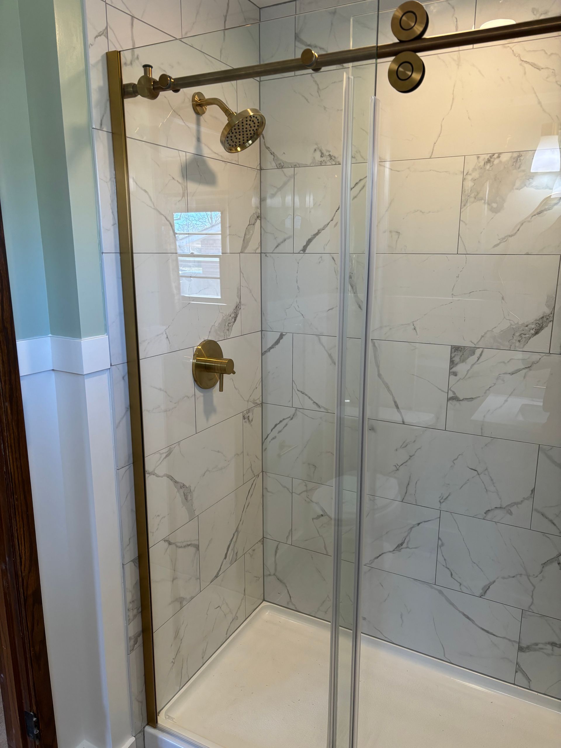 Shower with gold fixtures and marble-look tiles. Glass door slides on a track.