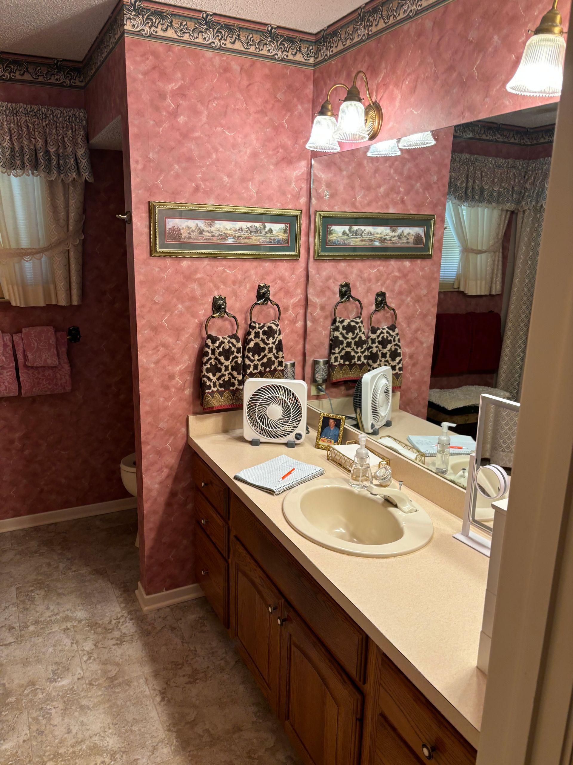 Bathroom with pink textured wallpaper, beige countertop, wooden cabinets, and large mirror.