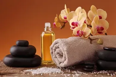 Spa setup with stacked black stones, rolled towel, oil bottle, orchid flowers, and sea salt on a wooden surface.