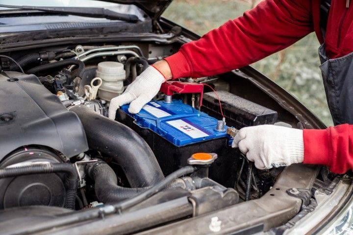 Person installing a blue car battery under the hood with white gloves and red sleeves