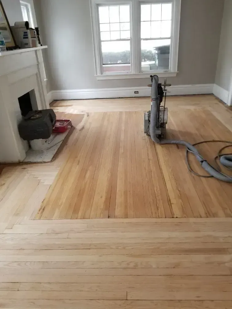 Wood floor being sanded in a living room near a fireplace and window.