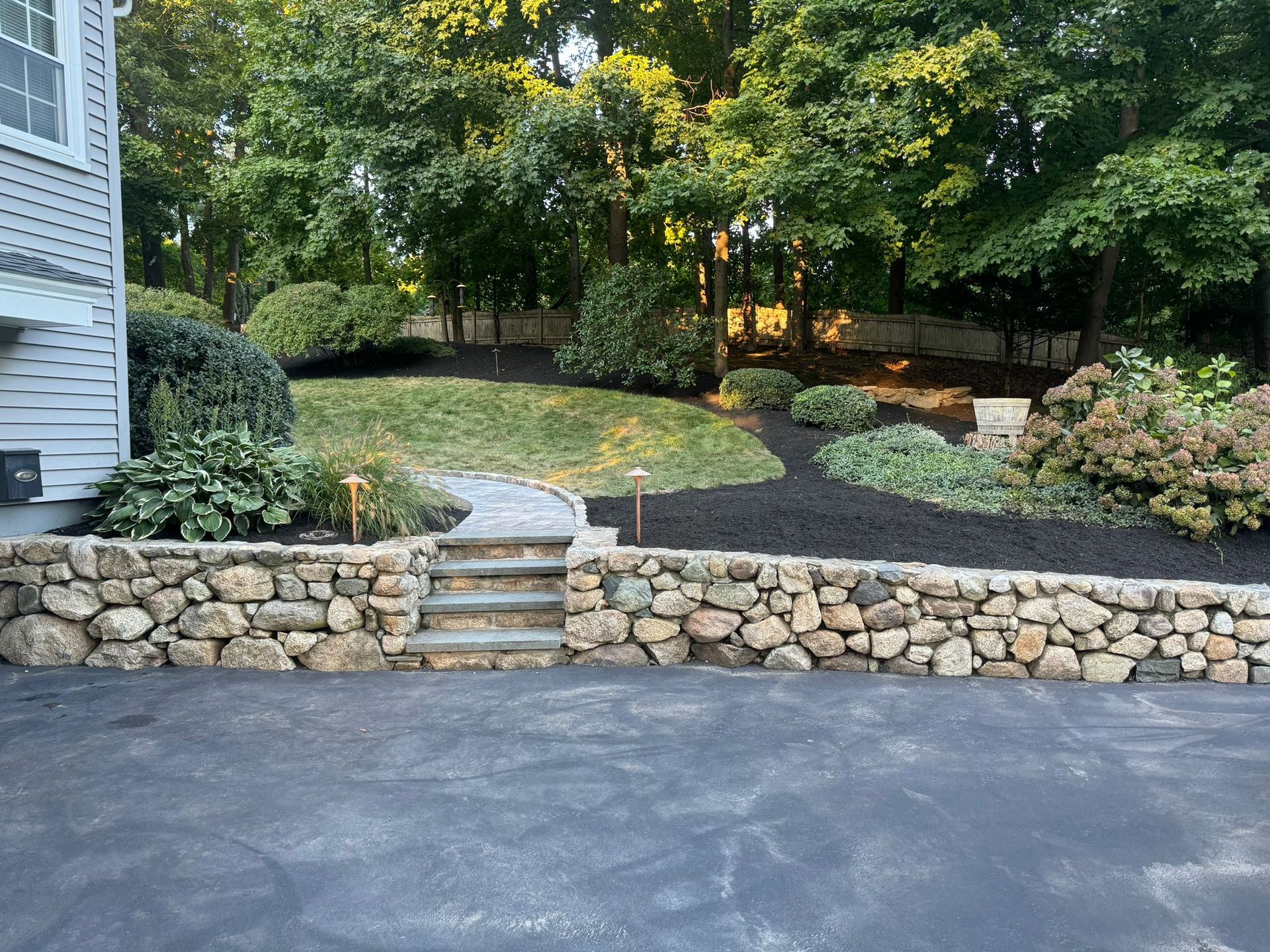 A stone wall with stairs leading up to a driveway in front of a house.