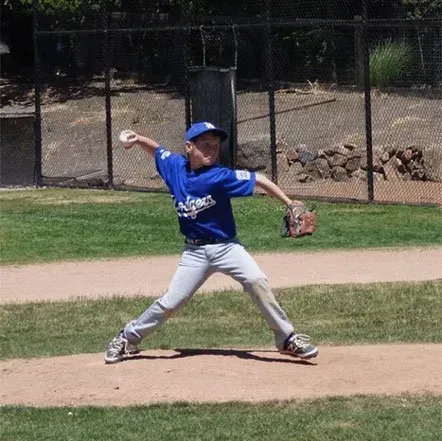 Young baseball player in blue uniform pitching on a dirt field, with a fence and rocks in the background