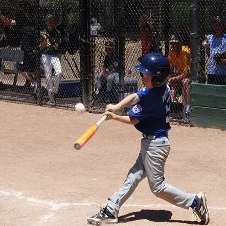 Young baseball player in blue swings an orange bat at a ball on a dirt field.