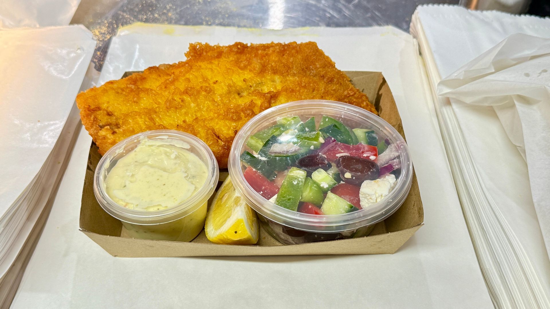 Fried fish, salad, sauce, and lemon in a takeout container on a counter.