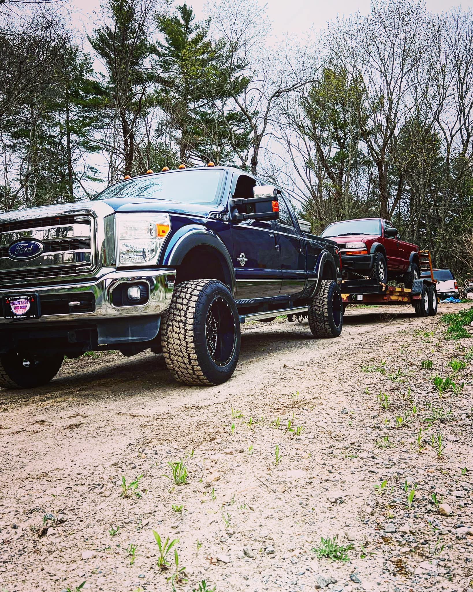 Dark blue lifted truck towing a red car on a trailer in a dirt area. Trees in the background.